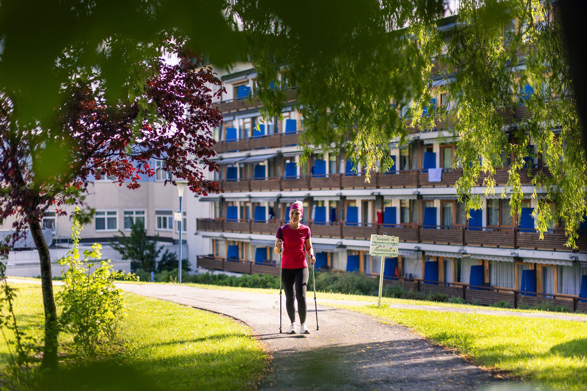 Eine sportlich gekleidete Frau mit Walking Stöcken trainiert auf einem Weg neben einem Hotel mit vielen Fenstern und Balkonen. Im Vordergrund ist erkennbar, dass der Weg durch Bäume und Sträucher schattig wird.