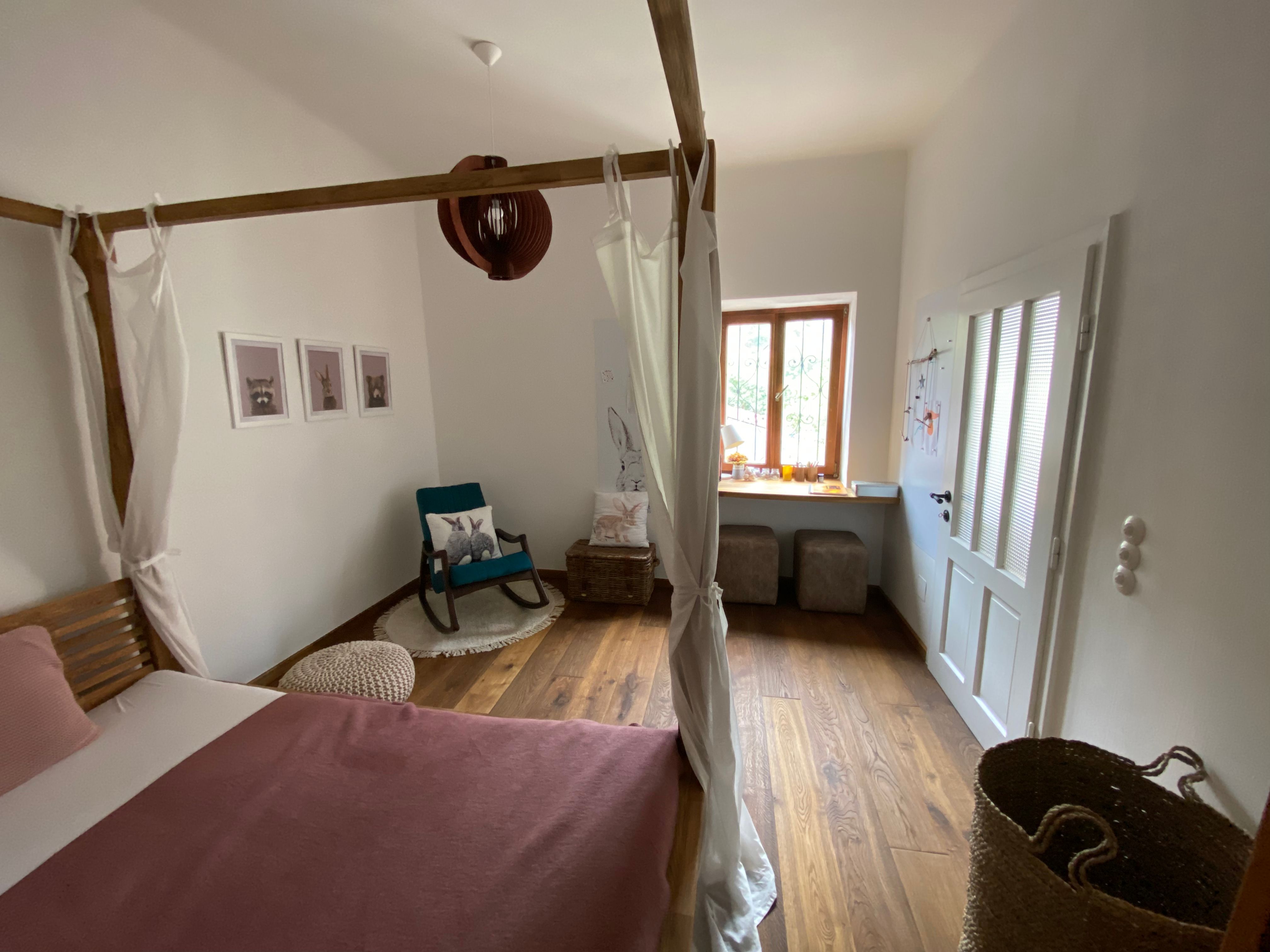 Gemütliches Schlafzimmer mit Himmelbett, Schaukelstuhl und Fensterbank. Holzboden und helle Wände.