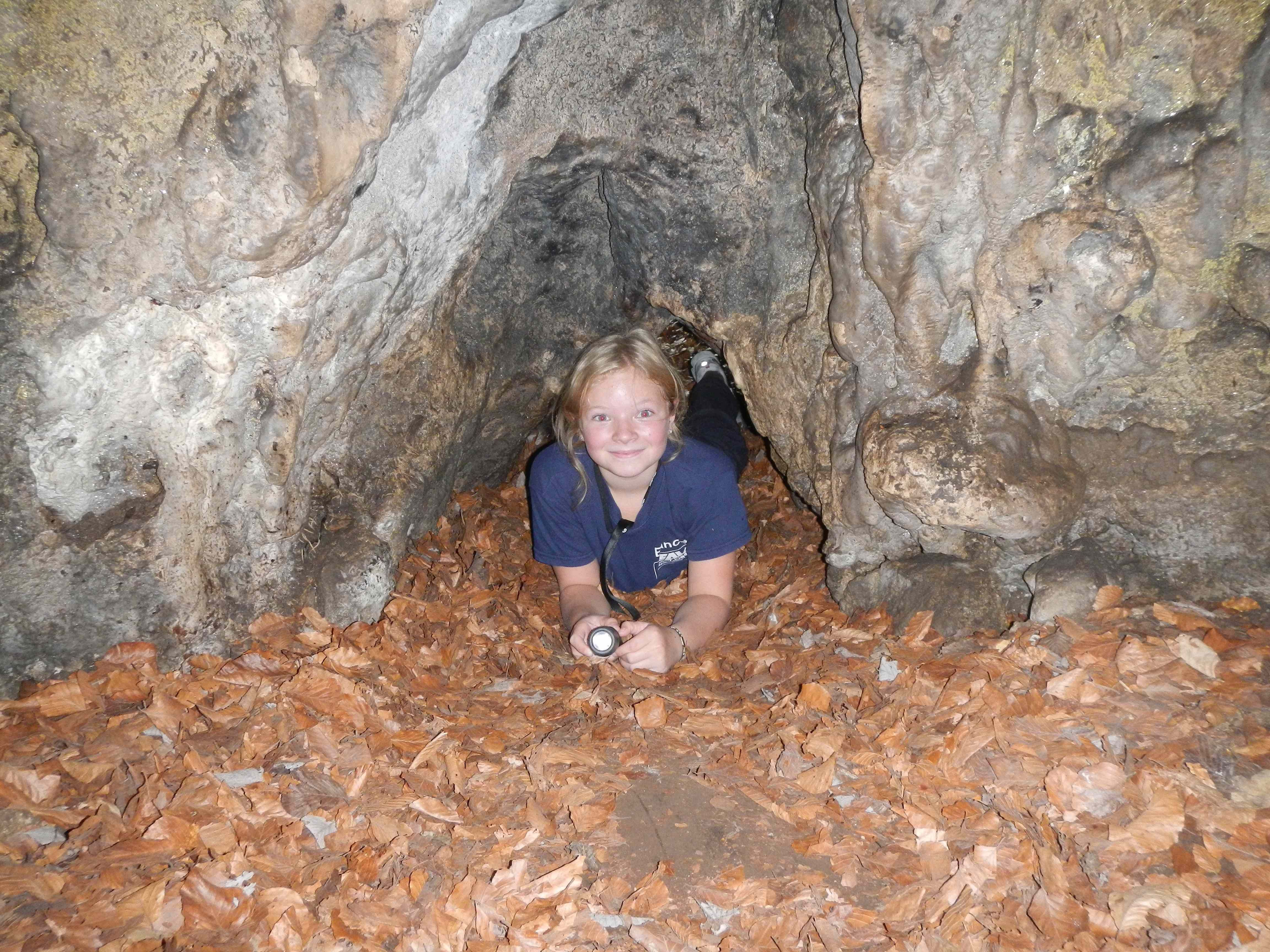 Ein Kind liegt in einer Höhle auf Laub und hält eine Taschenlampe.