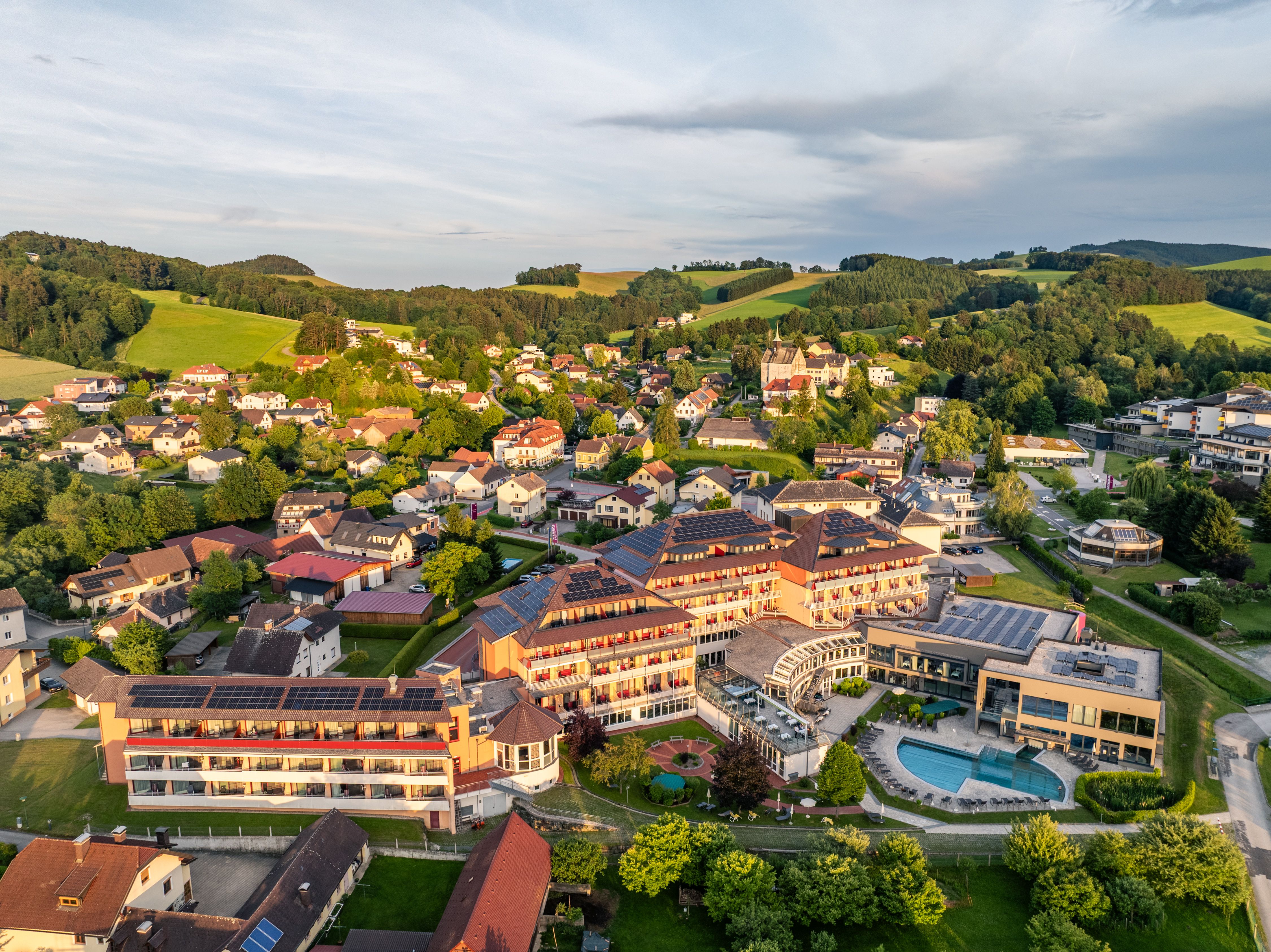 Luftaufnahme von Bad Schönau mit einem großen Hotel mit Pool im Vordergrund und grünen Hügeln im Hintergrund.