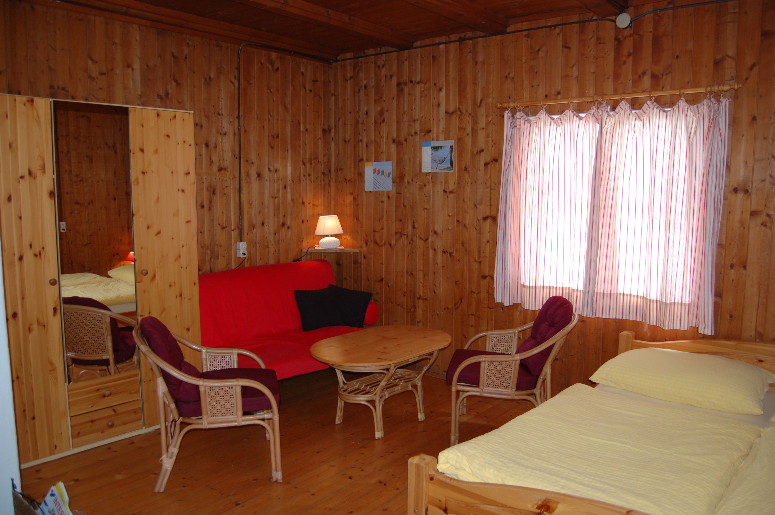 Innenansicht eines gemütlichen Zimmers mit Holzwänden, rotem Sofa, Tisch, Stühlen und Bett.