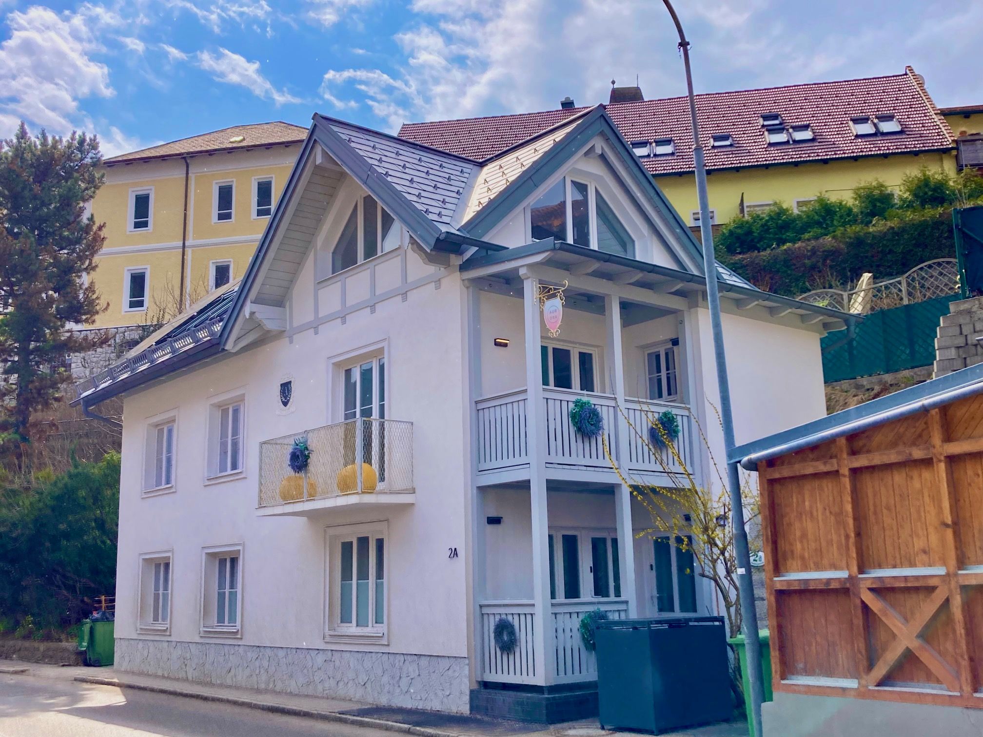 Ein modernes, weißes Haus mit blauen Dächern und einem kleinen Balkon, umgeben von weiteren Gebäuden und einem Baum.