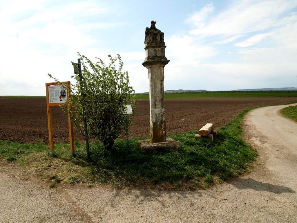 Steinsäule neben einem Feldweg mit Infotafel und Büschen.