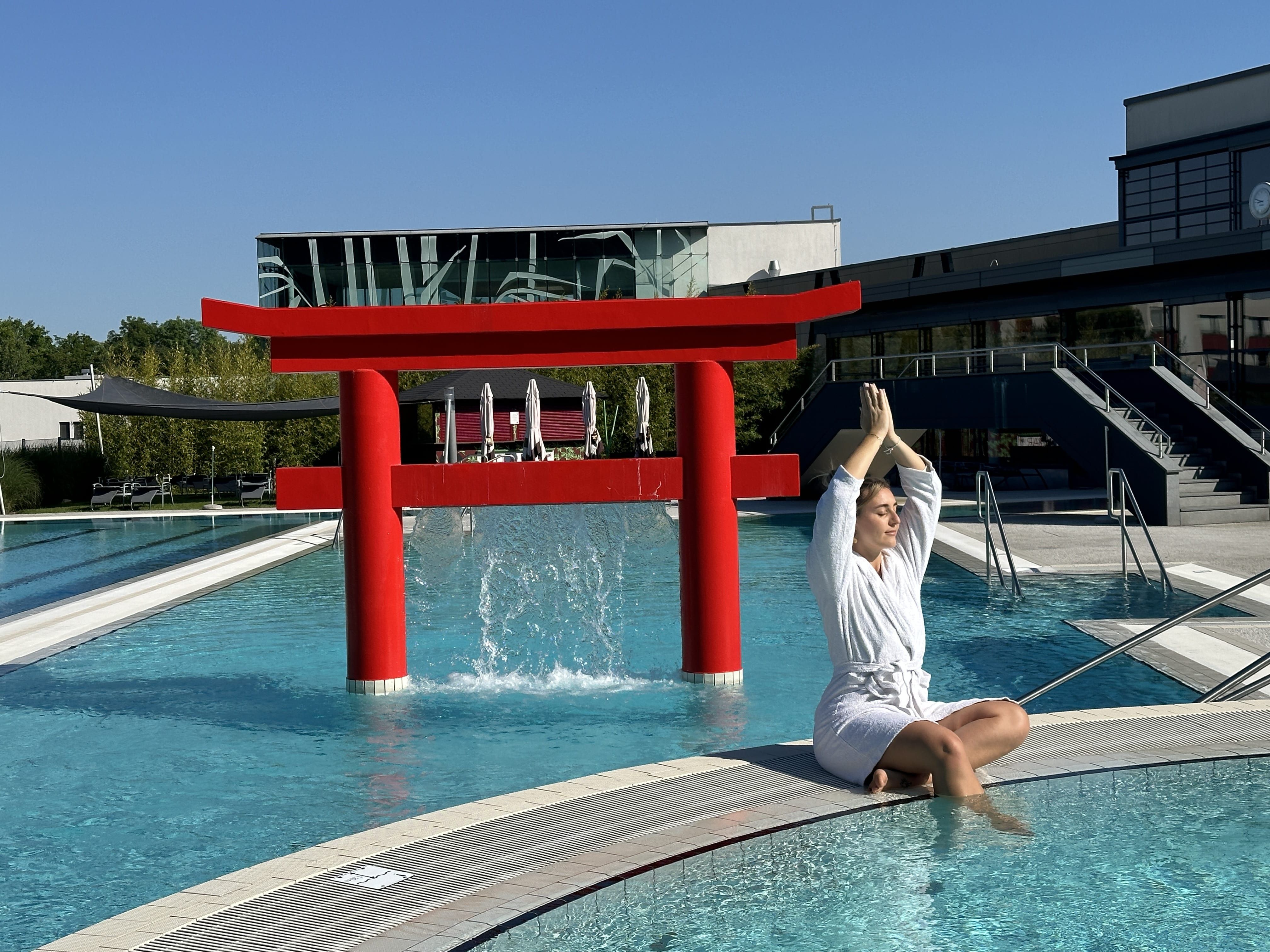 Eine Frau in einem weißen Bademantel macht Yoga am Rand eines Pools mit einem roten Torii im Hintergrund.