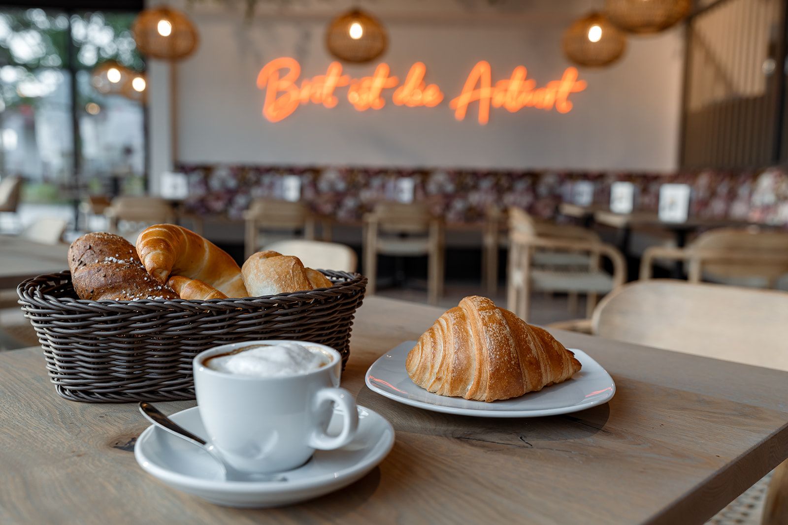 Ein Café mit einem Korb voller Gebäck, einem Croissant auf einem Teller und einer Tasse Cappuccino auf einem Tisch.