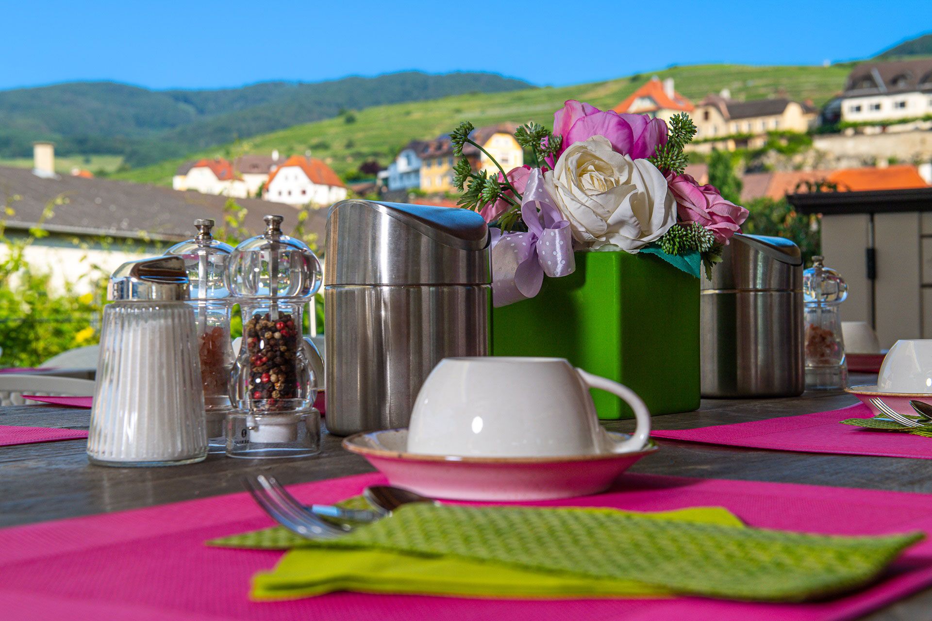 Frühstückstisch im Freien mit rosa Tischsets, Kaffeetassen, Salz- und Pfefferstreuern und Blumenarrangement, im Hintergrund eine malerische Landschaft.