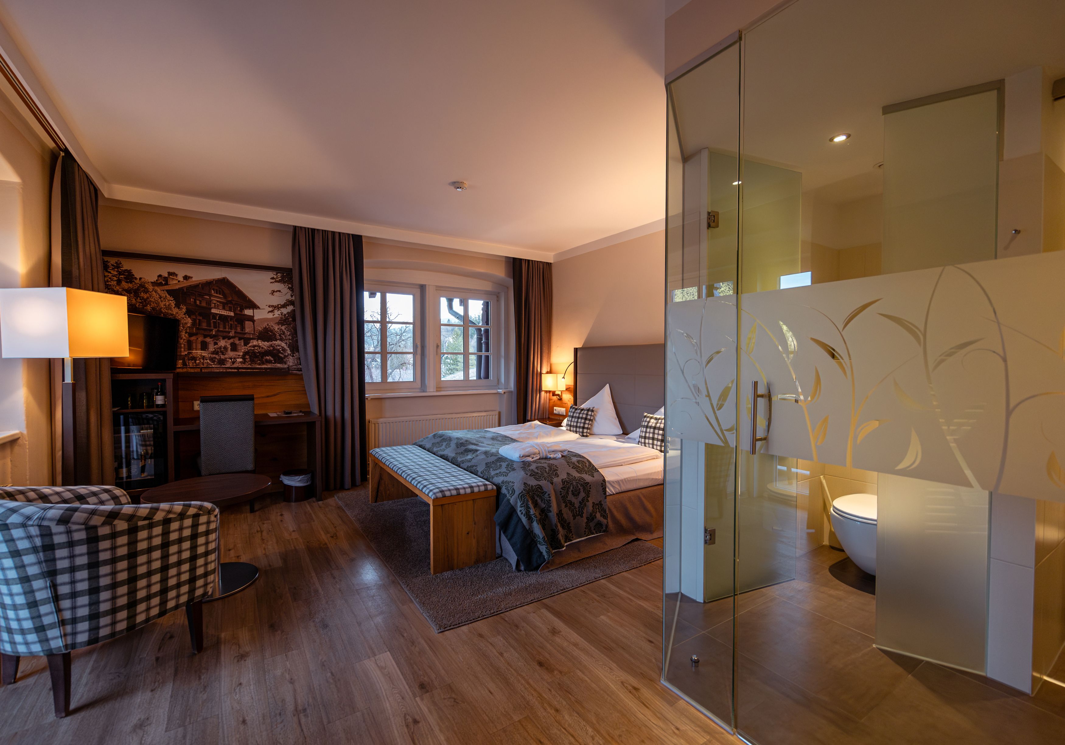 Hotelzimmer mit Doppelbett, Glaswand zum Bad, Sessel und Schreibtisch.