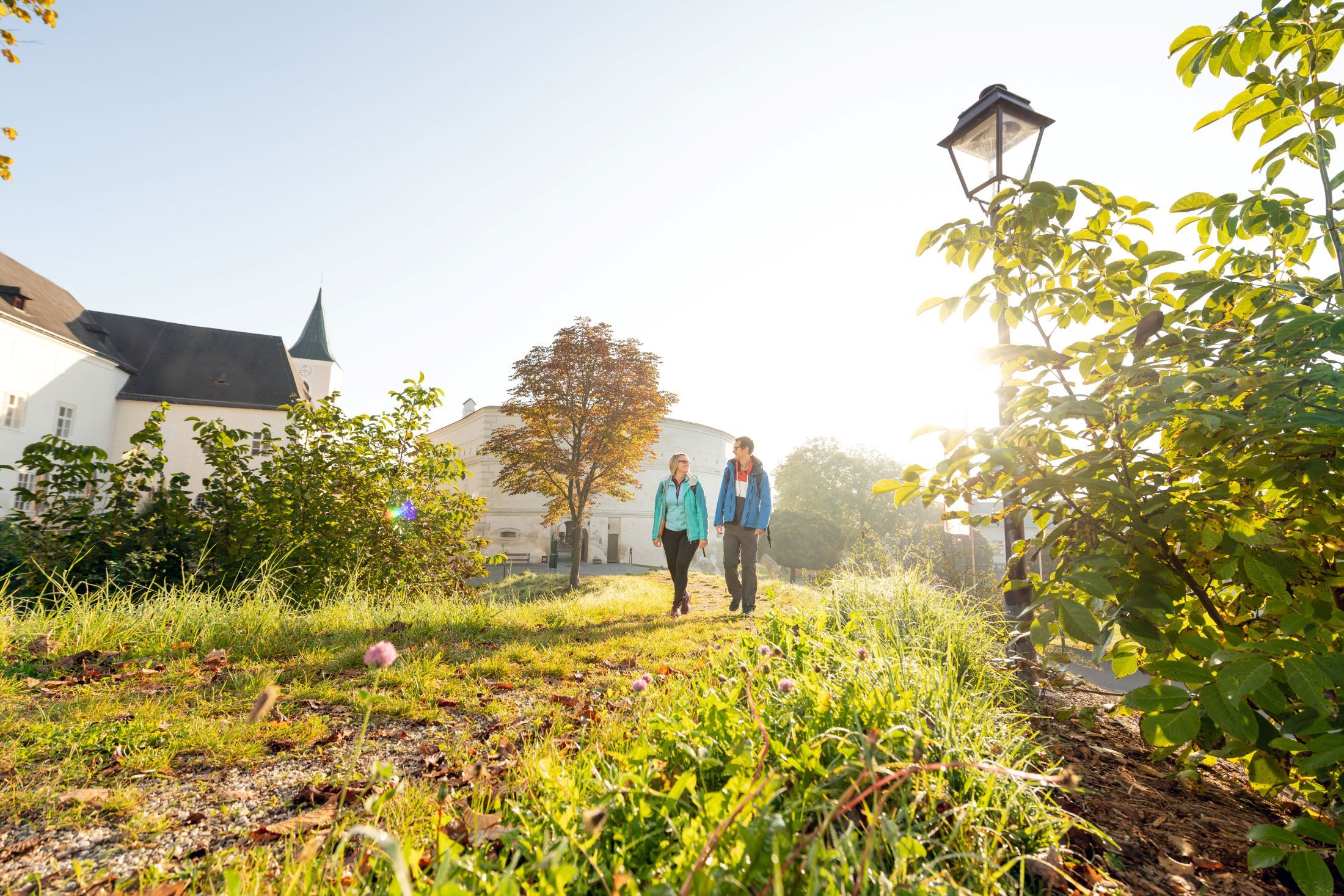 Zwei Personen spazieren auf einem sonnigen Weg neben einem Gebäude mit Turm.