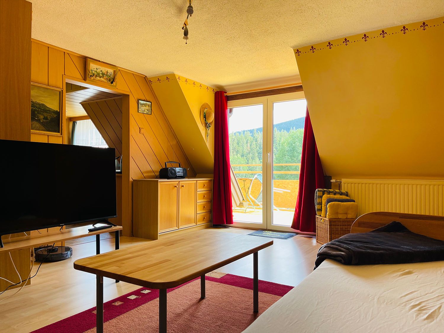 Gemütliches Wohnzimmer mit Holzmöbeln, großem Fernseher, Tisch und Zugang zu einem Balkon mit Aussicht auf grüne Landschaft.
