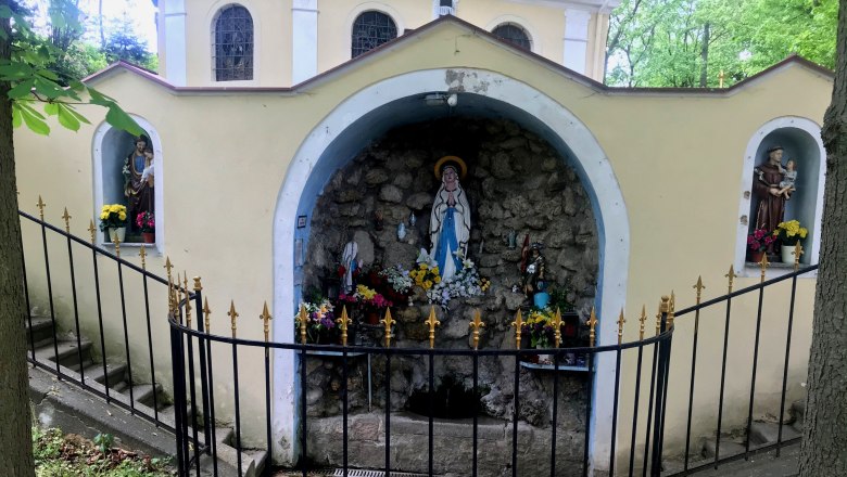 Eine Grotte mit religi&ouml;sen Statuen und Blumen, umgeben von einem schmiedeeisernen Zaun.