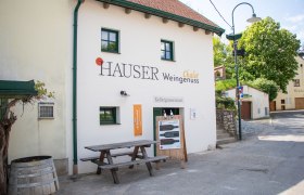 Außenansicht eines Gebäudes mit der Aufschrift 'Hauser Chalet Weingenuss'. Vor dem Gebäude steht ein Holztisch und ein Weinfass.