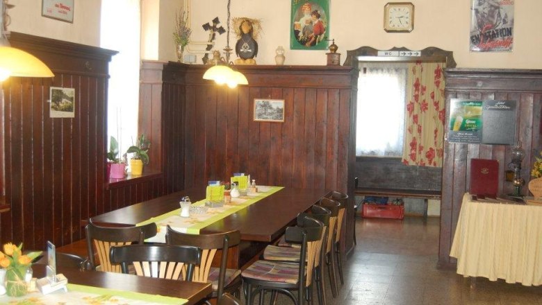 Gem&uuml;tlicher Gastraum mit Holzt&auml;felung, Tisch mit St&uuml;hlen, dekorativen Postern und einer Uhr an der Wand.