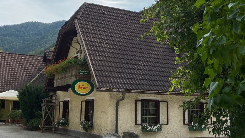 Traditionelles Gasthaus mit Blumen und Murauer-Schild, umgeben von Bäumen.
