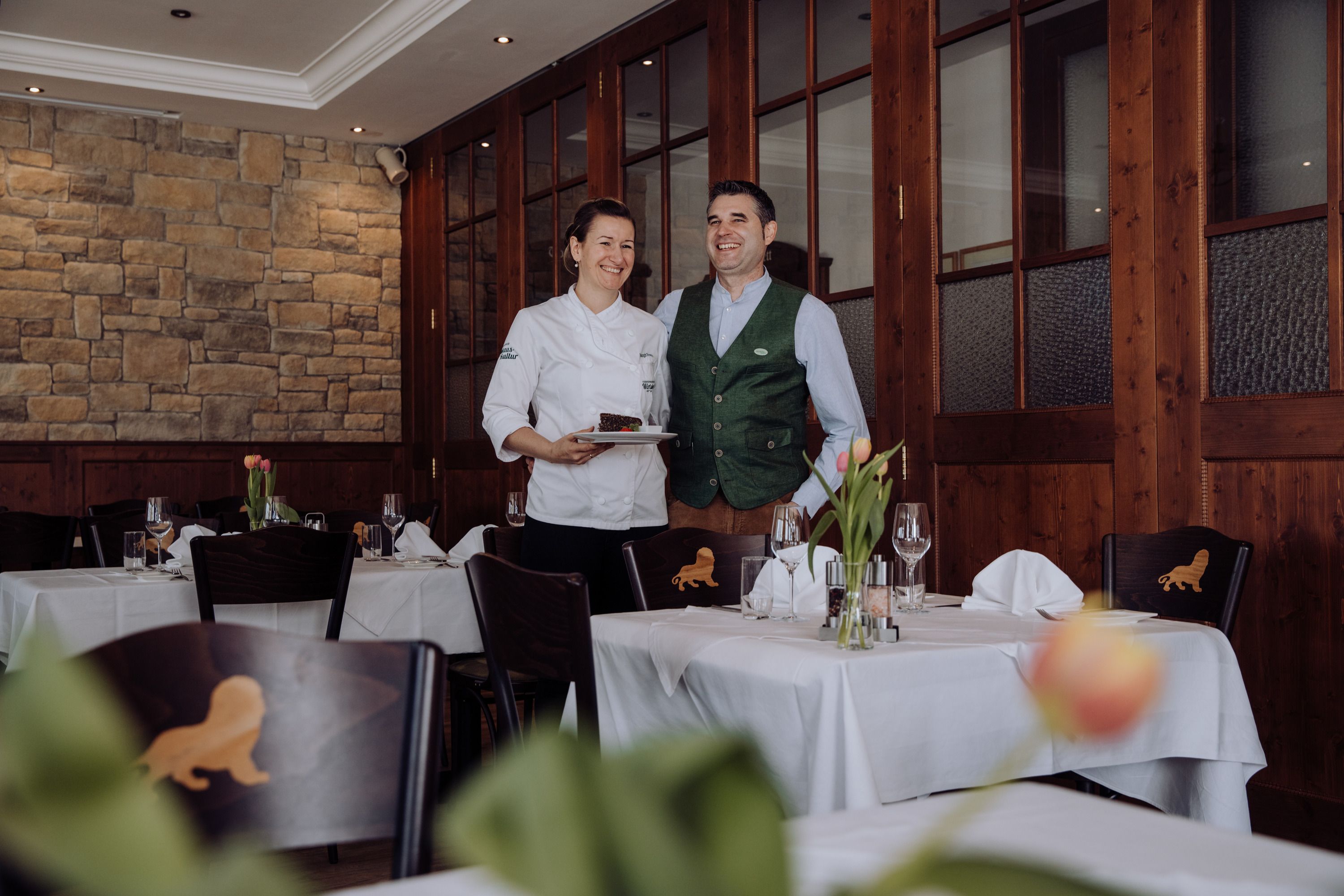 Eine Chefköchin und ein Mann in traditioneller Kleidung stehen lächelnd in einem eleganten Restaurant.