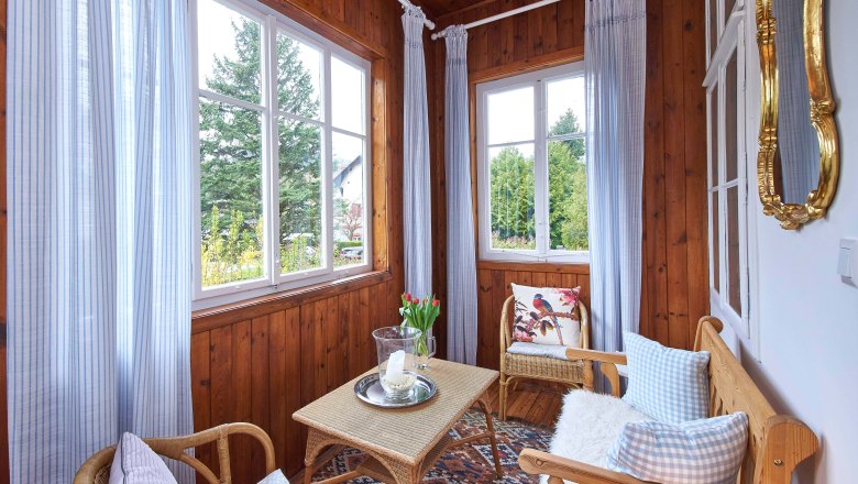 Ein gem&uuml;tlicher Wintergarten mit Holzw&auml;nden, Korbm&ouml;beln, blauen Vorh&auml;ngen und einem Tisch mit Kerze und Blumen.