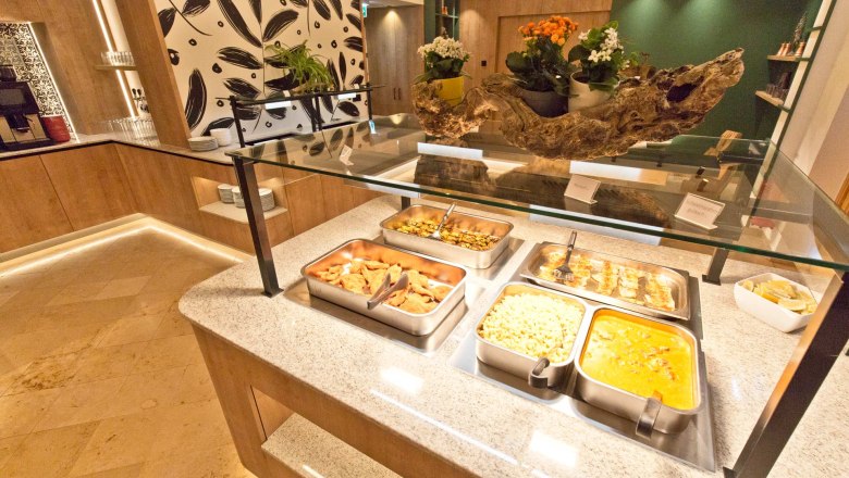 Buffetbereich mit verschiedenen Speisen in Edelstahlbeh&auml;ltern, dekoriert mit Pflanzen und Holzelementen.