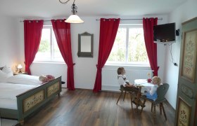 Ein traditionell eingerichtetes Zimmer mit Holzboden, roten Vorhängen und einem Tisch mit zwei Puppen in Tracht.