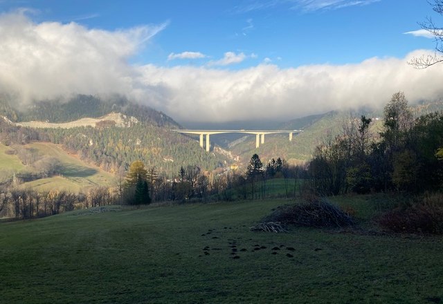 Landschaft mit Brücke über ein Tal, umgeben von bewaldeten Hügeln und Wolken.