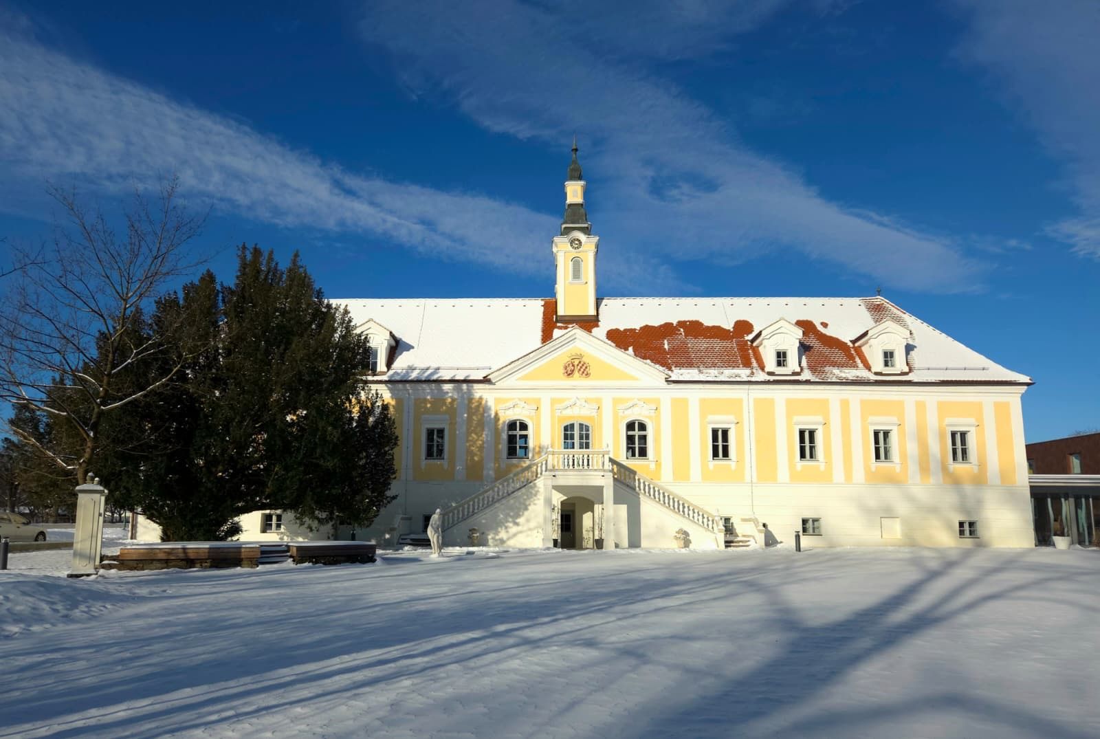 Winterbild von Hotel Schloss Haindorf: gelbes Schlossgebäude mit Turm, verschneitem Dach und Hof unter blauem Himmel.