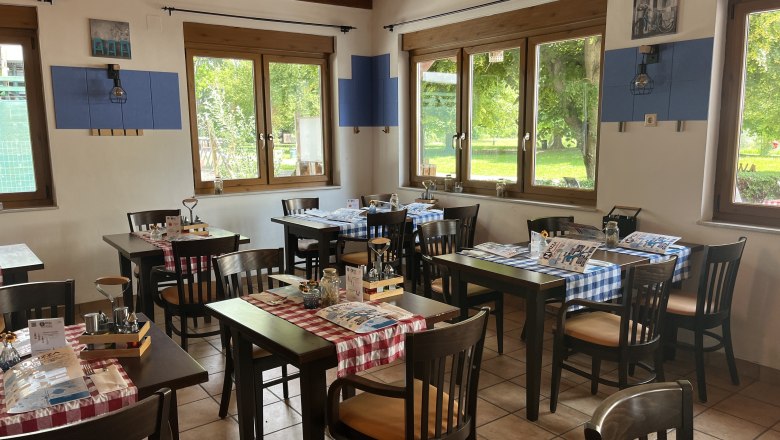 Innenansicht der Pizzeria. Schwarze M&ouml;bel mit roter Tischdecke und Besteck. Im Hintergrund ist durch die Fenster das Aubad zu sehen,