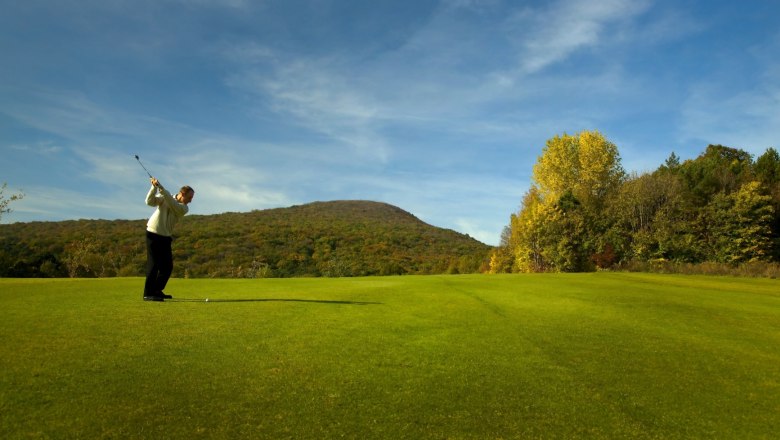 Golfer schlägt Ball auf einem grünen Golfplatz mit bewaldeten Hügeln im Hintergrund.