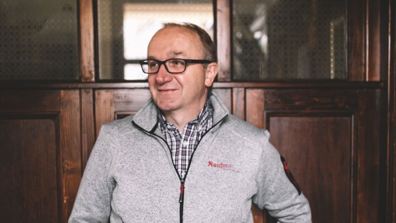 Ein Mann mit Brille und grauer Jacke steht vor einer Holzwand.