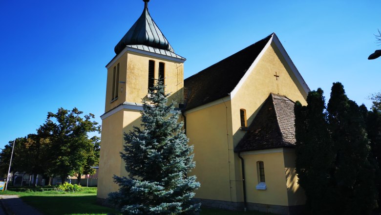 Gelbe Kapelle mit Turm und Kreuz, umgeben von B&auml;umen, unter blauem Himmel.