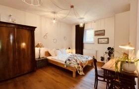 Gemütliches Schlafzimmer mit Holzboden, Doppelbett, antikem Schrank, Schreibtisch mit Lampe und Pflanze, sowie dekorativen Elementen an den Wänden.