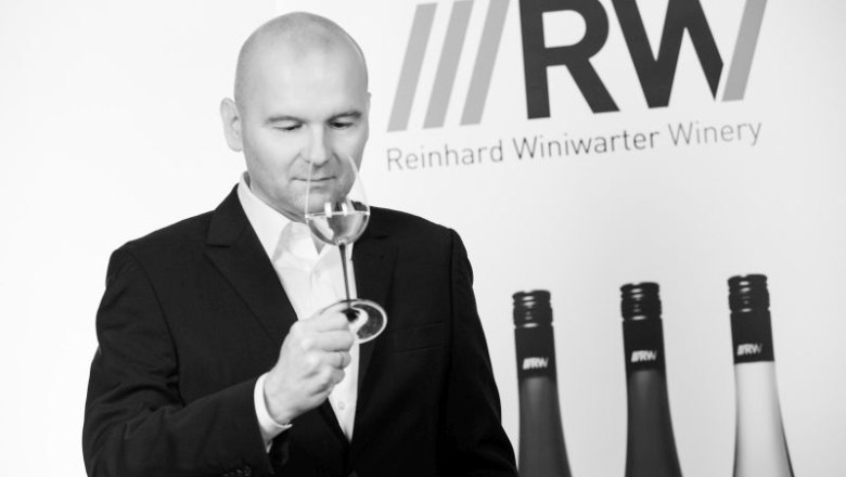 Mann in Anzug riecht an einem Weinglas vor einem Banner mit der Aufschrift 'Reinhard Winiwarter Winery'.