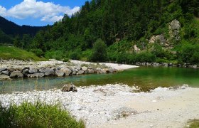 Flusslandschaft mit klarem Wasser, Kieselstrand und bewaldeten Hügeln.