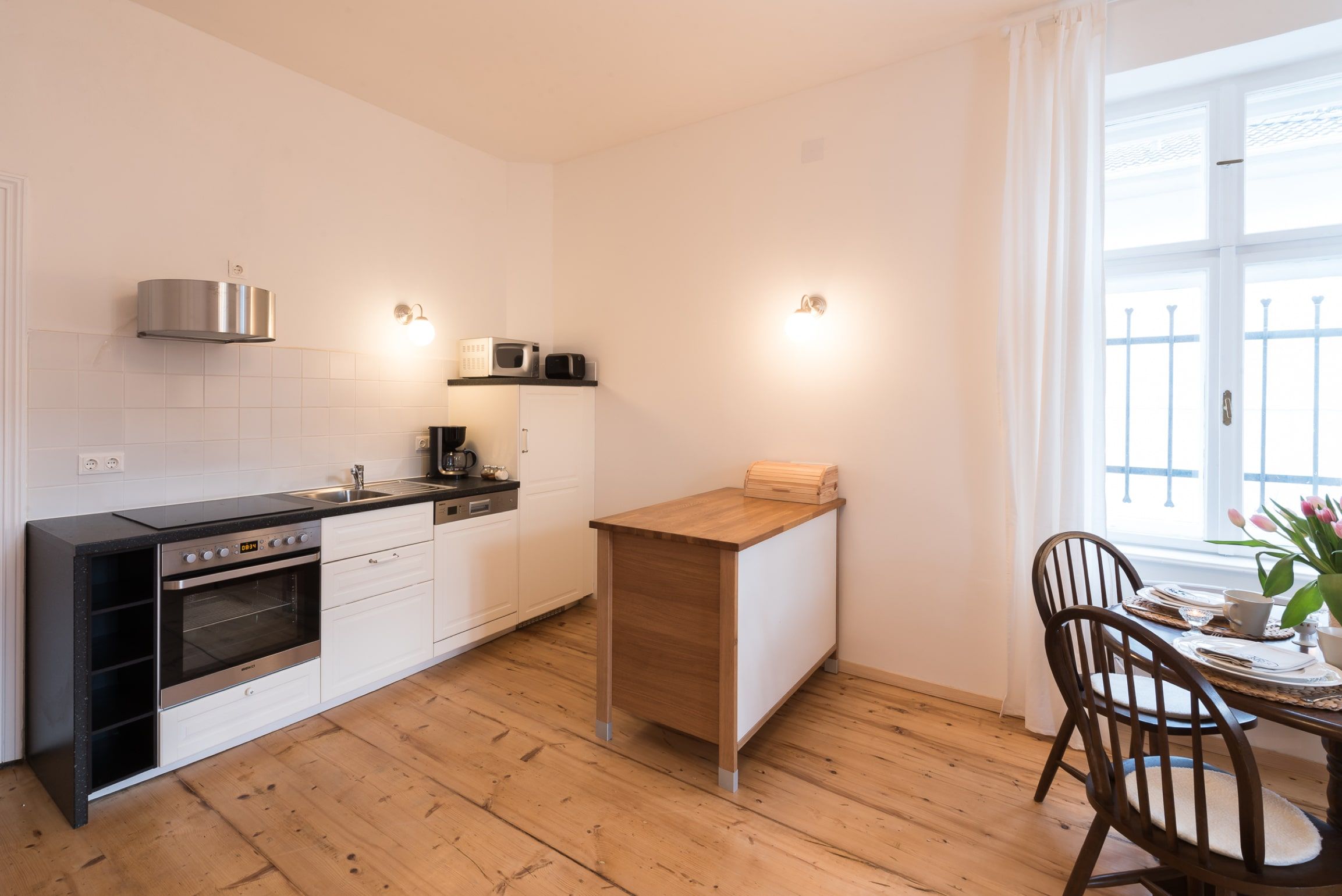 Moderne Küche mit Holzboden, weißer Einrichtung und kleinem Esstisch am Fenster.