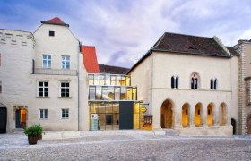 Historisches Gebäude mit modernem Glaszwischenbau, Kopfsteinpflaster im Vordergrund.