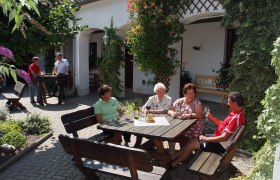 Menschen sitzen an einem Tisch im Freien und genießen Getränke in einem begrünten Innenhof.