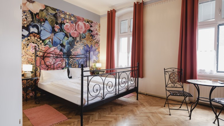 Ein stilvoll eingerichtetes Zimmer mit einem schmiedeeisernen Bett, floraler Wandtapete und einem kleinen Tisch mit Stühlen vor großen Fenstern.