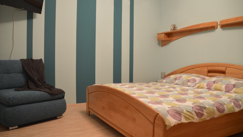 Schlafzimmer, © Eva Stuhr Schlafzimmer mit gestreifter Wand, Holzbett und Sofa.