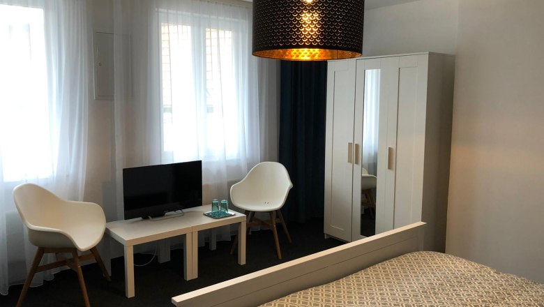 Modern eingerichtetes Doppelzimmer mit Bett, Schrank, zwei St&uuml;hlen, kleinem Tisch und Fernseher.