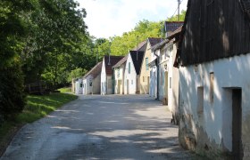 Reihe von Weinkellern in einer l&auml;ndlichen Stra&szlig;e mit B&auml;umen im Hintergrund.