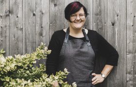 Eine Frau mit Brille und Schürze steht lächelnd vor einer Holzwand neben einem Busch.