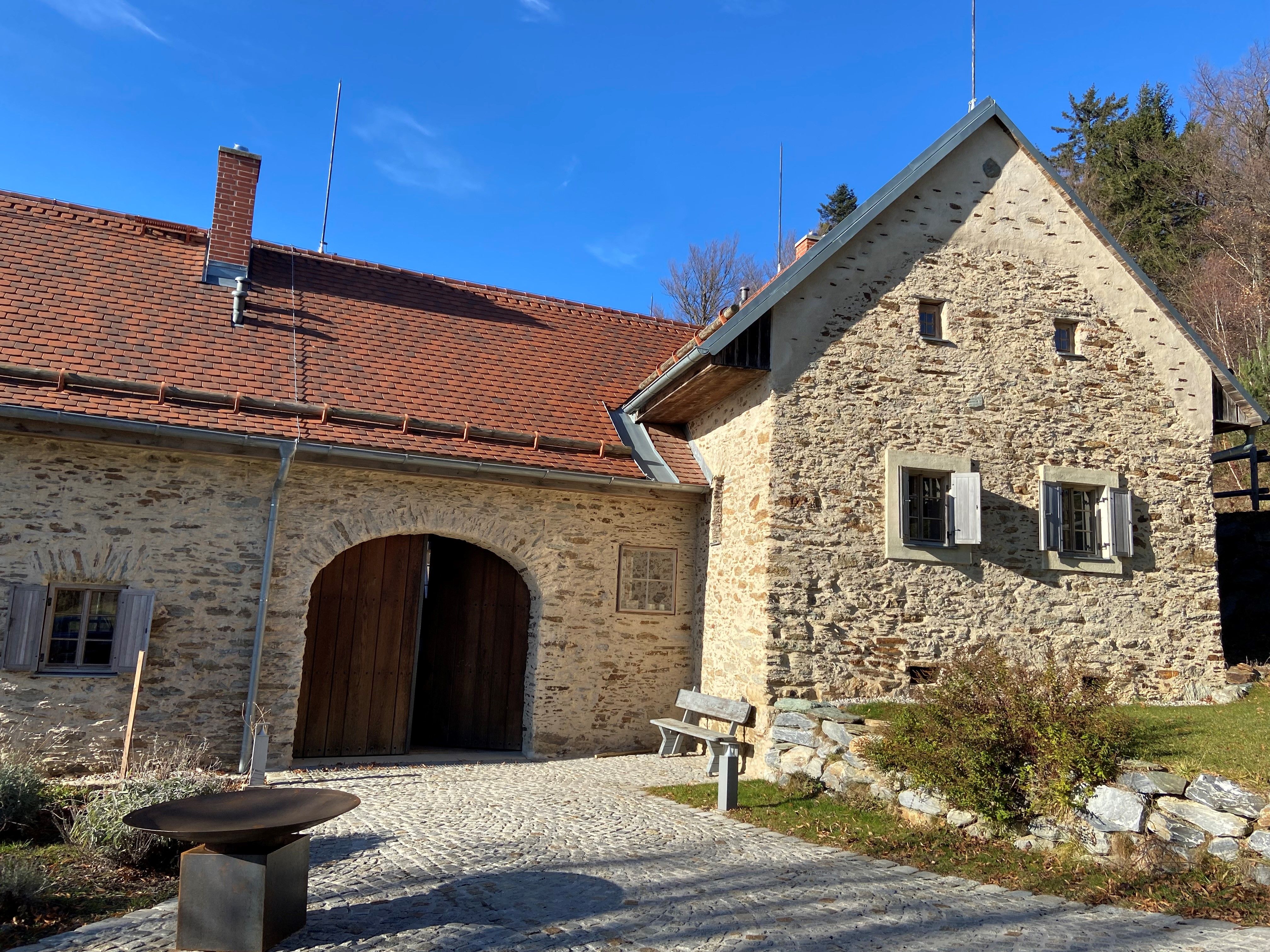 Altes Steingebäude mit rotem Ziegeldach und Holztor, umgeben von gepflastertem Weg und Vegetation.