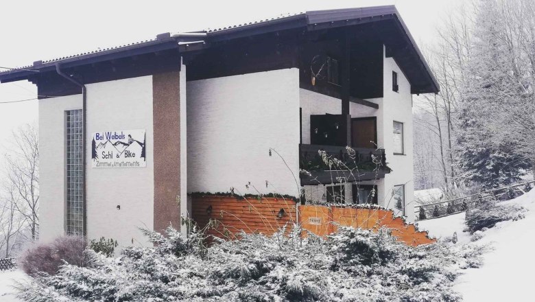 Ein verschneites Gebäude mit einem Schild 'Bei Webels Ski & Bike Zimmer-Apartments'.