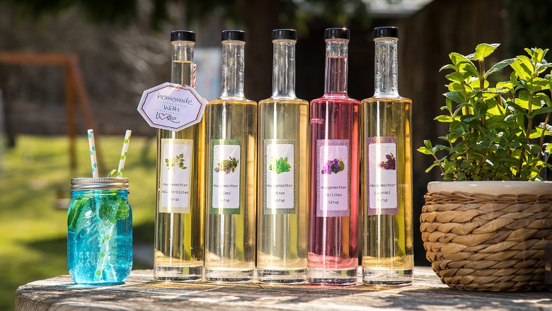 Fünf Flaschen mit selbstgemachtem Sirup und ein blaues Glas mit Strohhalmen auf einem Holztisch im Freien.