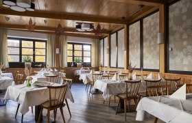 Ein elegantes Restaurant mit Holzm&ouml;beln, wei&szlig;en Tischdecken und gro&szlig;en Fenstern, die herbstliche B&auml;ume zeigen.