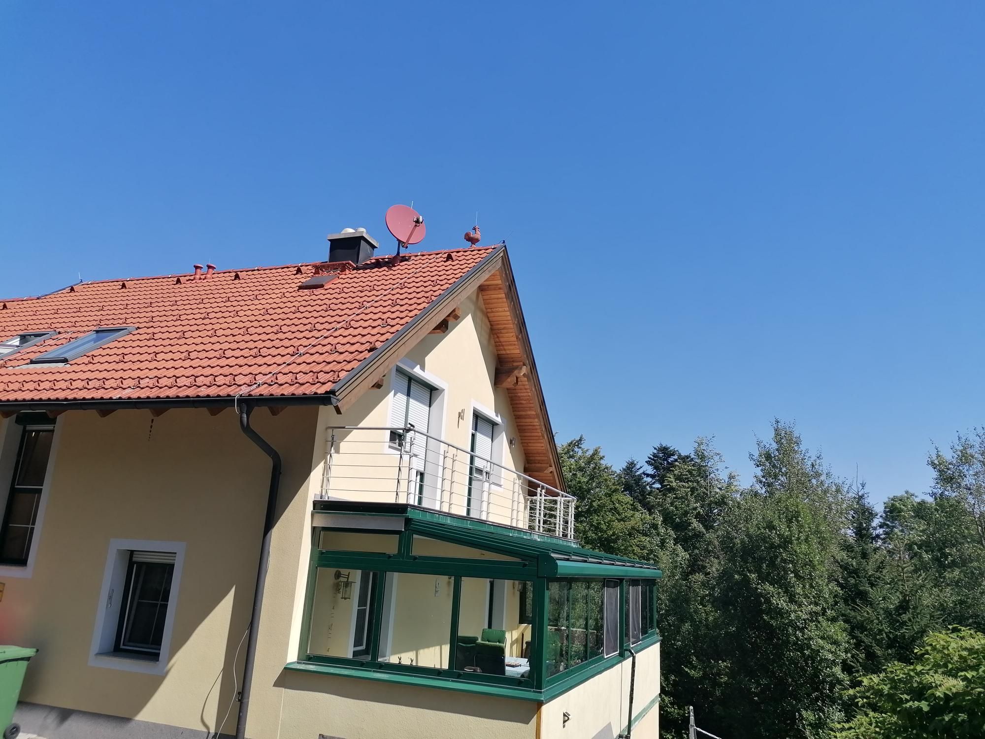 Gelbes Haus mit rotem Dach und Balkon, umgeben von Bäumen, unter blauem Himmel.