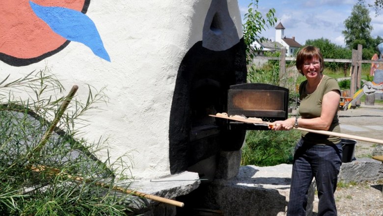 Eine Frau steht vor einem bemalten Ofen im Freien und h&auml;lt einen langen Holzschieber.