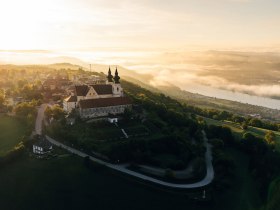 Die sanften H&uuml;gel umgeben die majest&auml;tische Basilika, w&auml;hrend die Donau in der Ferne glitzert. Ein atemberaubender Sonnenaufgang taucht die Landschaft in warmes Licht und l&auml;dt Wanderer ein, die Sch&ouml;nheit der Natur zu genie&szlig;en. Hier, wo der Himmel die Erde k&uuml;sst, entfaltet sich ein wahres Paradies der Blicke.