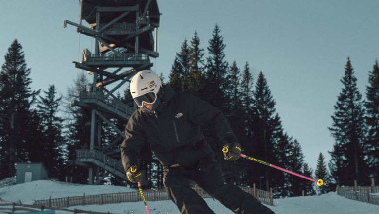 Skifahrer in schwarzer Kleidung f&auml;hrt auf pr&auml;parierter Piste am Semmering Hirschenkogel, im Hintergrund ein Aussichtsturm und B&auml;ume.