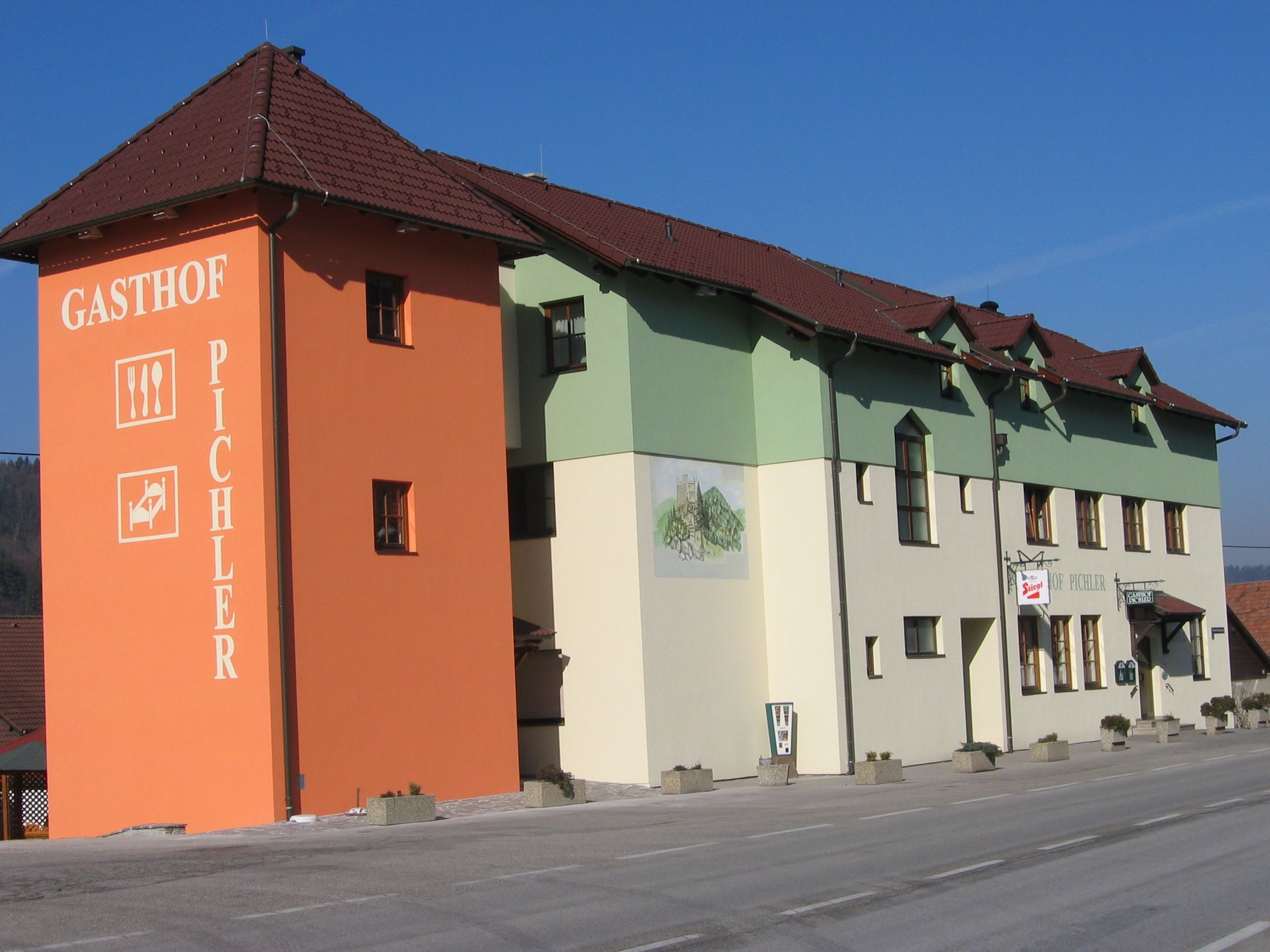 Ein Gasthof mit orangefarbenem Turm und grün-weißer Fassade an einer Straße.