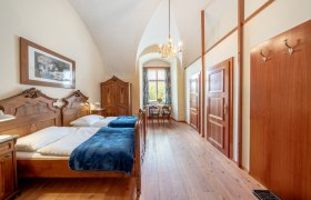 Ein elegantes Doppelzimmer mit antiken Holzm&ouml;beln, einem Kronleuchter und einem Fenster mit Blick ins Gr&uuml;ne.