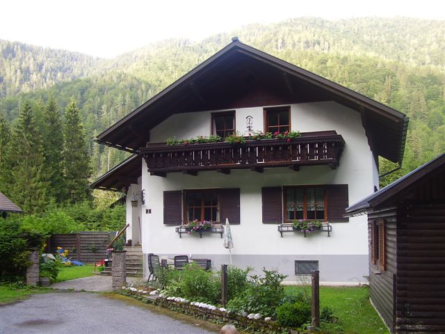 Ein traditionelles Ferienhaus mit Holzbalkon und Blumenkästen, umgeben von grünen Bäumen und Bergen.