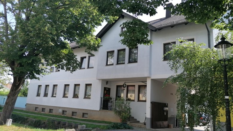 Au&szlig;enansicht eines wei&szlig;en Geb&auml;udes mit vielen Fenstern, umgeben von B&auml;umen.